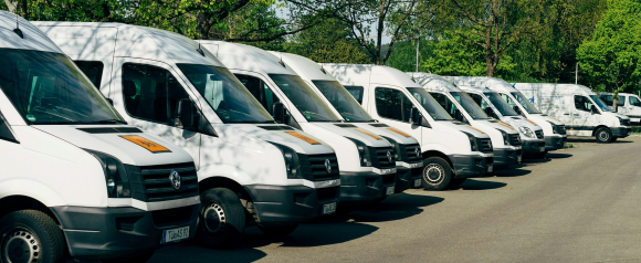 A line of white commercial vans parked side by side under leafy trees in a sunny lot, suggesting organization and readiness for dispatch. The image reflects how Fleetr’s fleet tracking system helps manage vehicle operations efficiently and in real time.