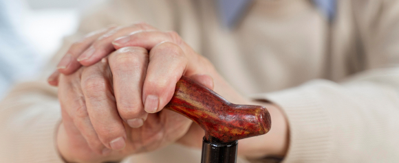 Close-up of an elderly person's hands resting on a wooden cane, wearing a beige sweater. The image conveys themes of aging, mobility assistance, and the importance of support systems like medical alerts or Medical Guardian devices.