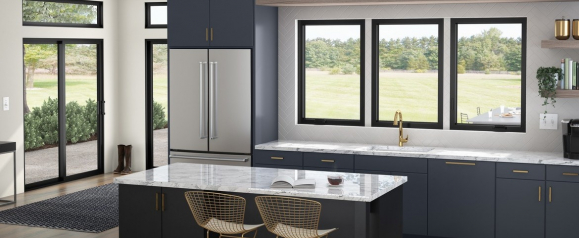 A modern kitchen with sleek black-framed Milgard windows that provide a clear view of a sunny backyard and fill the space with natural light. The minimalist window design pairs well with dark cabinetry, gold hardware, and marble countertops for a contemporary look.