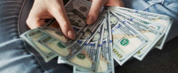 Close-up of a person holding a fan of $100 bills while sitting in jeans, symbolizing financial freedom or a recent cash loan. The image conveys the potential benefits of Achieve personal loans for quick access to funds.