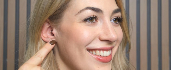 A blonde woman smiling and pointing to the discreet Soundbright hearing aid fitted inside her ear canal.