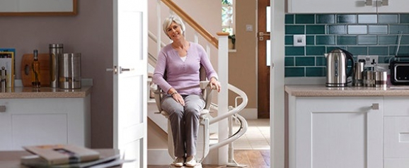 An older woman with short gray hair rides a curved stairlift down into a modern kitchen, smiling and appearing at ease. The image highlights how Stannah stairlifts provide safe, stylish mobility solutions for navigating multi-level homes.
