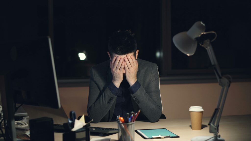A stressed employee sitting at a desk late at night with hands over their face, illustrating severe workplace distress that may require consulting an employment lawyer near me.