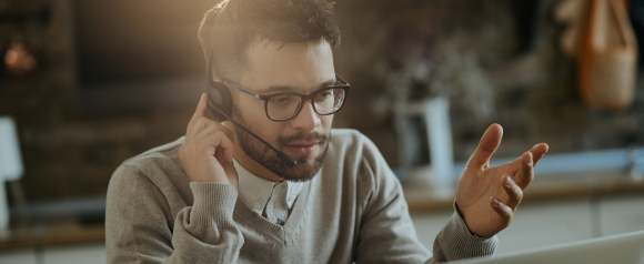 A man wearing glasses and a headset gestures with his hand while speaking, appearing focused during a remote video call. The setting is a home or casual office, highlighting the flexibility and clarity of VoIP-based business phone systems like Vonage.