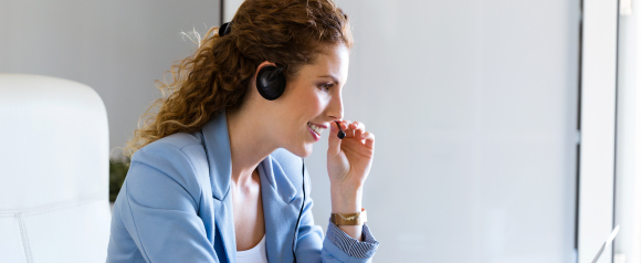 Smiling woman in a light blue blazer using a headset while speaking to a customer, seated at a desk in a bright office. Represents professional customer service or sales support for 800.com’s VoIP business phone systems.