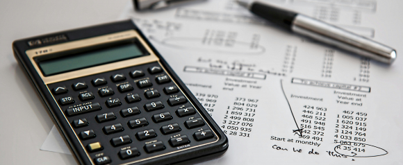Close up of a financial calculator resting on printed investment and expense reports with a pen placed on top of the documents. The papers show columns of numbers, projected values, and handwritten notes, emphasizing detailed financial analysis and budgeting. The image represents how NetSuite accounting software helps streamline reporting, track investments, and manage business finances accurately.