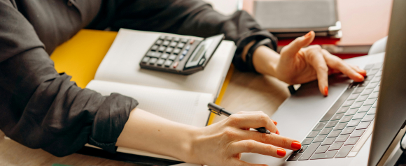 Close up of a person working on a laptop while holding a pen, with a calculator resting on an open notebook beside them. The desk setup highlights managing expenses, tracking numbers, and handling daily bookkeeping tasks. The image represents how FreshBooks accounting software helps streamline invoicing, organize finances, and simplify small business accounting.