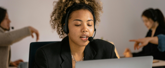 Woman using VoIP headset at laptop in a busy office, representing Aircall business phone systems.