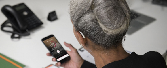 A woman with gray hair in a neat bun sits at a desk and looks at her smartphone, which displays a hearing aid control app interface. She wears a sleek, modern hearing aid in her right ear, possibly from Amplifon, and appears to be adjusting settings digitally.
