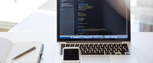 Open laptop on a clean desk displaying lines of computer code on the screen with a notebook, pen, and smartphone placed beside it, representing everyday online work and development. The image highlights the importance of antivirus protection like Norton to help safeguard devices and sensitive data from cyber threats while working online.
