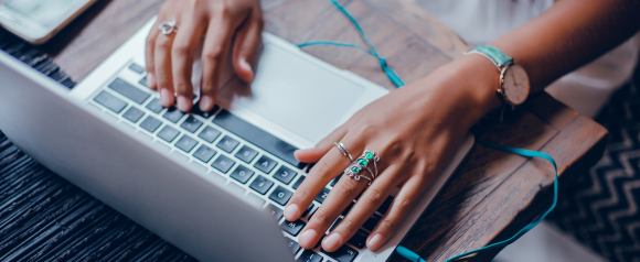 Close up of hands wearing silver and green gemstone rings typing on a laptop keyboard with teal earphones resting nearby on a wooden desk. The image represents everyday online activity and highlights the importance of Avast antivirus protection to help keep personal data and devices secure while browsing or working.