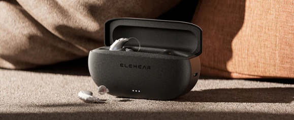 A sleek black Elehear hearing aids charging case sits open on a beige fabric sofa with one hearing aid inside and another resting beside it. The Elehear logo is visible on the front of the case, highlighting the modern design and portability of these hearing aids for everyday use.