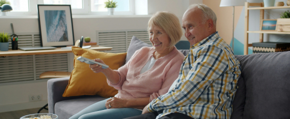 Smiling senior couple sitting on the couch watching TV with popcorn, representing the comfort and peace of mind offered by Galaxy Medical Alert Systems and medical alerts.