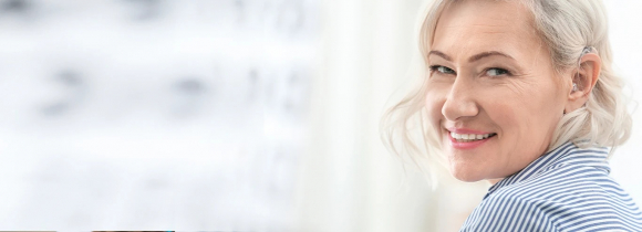 Smiling older woman with short blonde hair wearing a small behind the ear hearing aid with a clear tube, turning toward the camera in a bright indoor setting. The close up shows how modern hearing aids fit comfortably and discreetly along the ear. This image highlights the sleek design and everyday wearability of hearing aids available through hear.com.