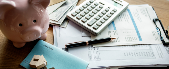 A piggy bank sits beside a calculator, cash, invoices, and a blue cashbook labeled cashbook on a wooden desk, with a set of house keys and a small wooden house placed on top. The scene represents budgeting and comparing personal loans through LendingTree for home related expenses or financial planning.