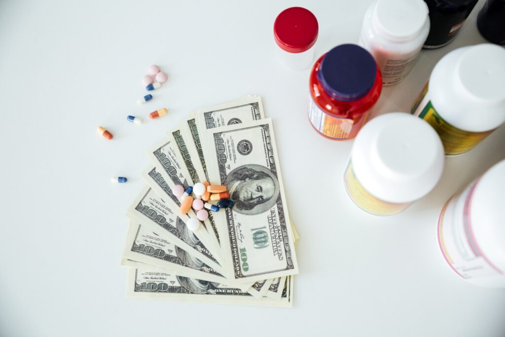 Stack of one hundred dollar bills with assorted colorful pills scattered on top beside several prescription bottles on a white surface. The image highlights the high cost of medication and the financial burden that leads many people to seek medical debt relief.