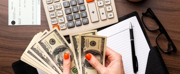 Hands counting a stack of hundred-dollar bills next to a paycheck, calculator, and planner on a desk, representing payroll management with Homebase.