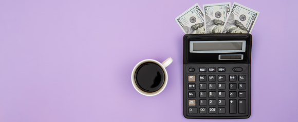 Black calculator with several one hundred dollar bills tucked into the top sits beside a cup of black coffee on a purple background. The image represents managing business finances and payroll expenses with Patriot Software payroll tools. The simple layout highlights budgeting, cash flow, and accurate payroll calculations for small businesses.