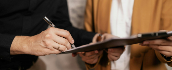A close up of a person signing paperwork on a clipboard while another person in a blazer holds the documents steady, suggesting an in person meeting about personal loans through LendingClub. The focus on the pen and contract highlights the loan application or approval process.