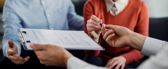 A person holds out a clipboard with loan documents while offering a pen for signature as a couple reviews the paperwork together. The image reflects the application process for Truist personal loans, highlighting an in person meeting to finalize financial paperwork.