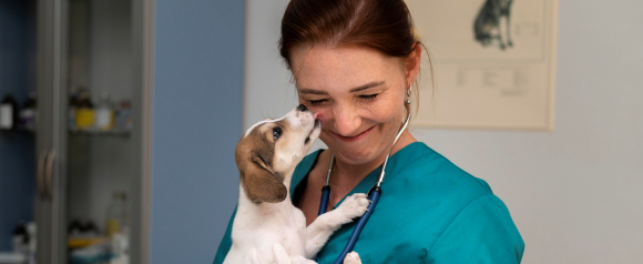 Veterinarian in teal scrubs with a stethoscope smiles as a small puppy reaches up to lick her nose in a bright exam room. The image highlights the caring bond between pets and providers, reinforcing how pet insurance from Trupanion helps support unexpected veterinary visits and protect a pet’s health.