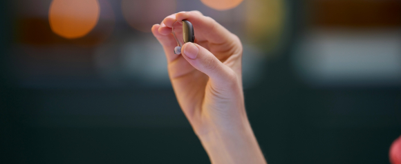Close up of a hand holding a small behind the ear ReSound hearing aid between the fingers, showing the slim curved design and soft ear tip against a softly blurred background. The image highlights how ReSound hearing aids are discreet and comfortable, emphasizing their compact size and modern design for everyday wear.