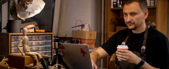 A small business owner sits at a workbench with a laptop and takeaway coffee, surrounded by organized drawers of tools and supplies in a cozy workshop. The image reflects the daily operations of an entrepreneur managing tasks online, highlighting the need for reliable digital protection like Norton Small Business.