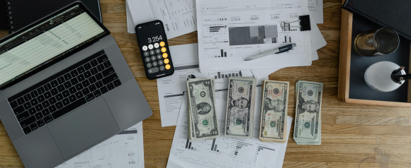 Desk workspace with a laptop, printed financial reports, a smartphone calculator displaying 3,254, and organized stacks of U.S. dollar bills. The setup suggests payroll processing or business accounting tasks, illustrating a real-world use case for Square Payroll.