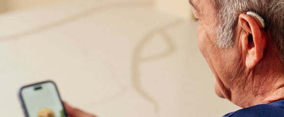 Side view of a senior man wearing a behind the ear hearing aid while using a smartphone at home. The sleek device rests comfortably behind his ear, highlighting modern design and everyday usability. The image showcases Starkey hearing aids as a smart solution for staying connected and improving daily hearing clarity.