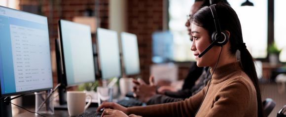 Customer service representative wearing a headset works at a computer in a modern office, speaking with a client while viewing a call management dashboard on screen. The image highlights how Net2Phone voip supports Business Phone Systems by enabling efficient communication and real time customer support in a professional workspace.