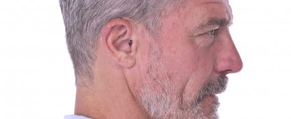 Side profile of an older man with short gray hair wearing a discreet in ear hearing aid with a thin clear wire that curves behind his ear. The close up highlights how modern hearing aids fit comfortably and blend naturally with the ear for everyday use. This image reflects the subtle design options available through ZipHearing for those seeking nearly invisible hearing aids.