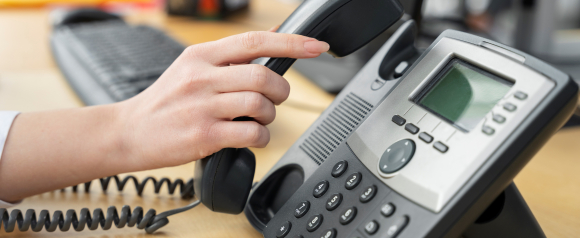 Hand lifting the receiver of an office desk phone on a work desk, illustrating a business communication setup powered by Nextiva business phone systems and VoIP technology.