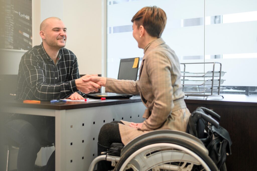 Man in office shaking hands with a woman in a wheelchair across a desk with paperwork and a laptop during a meeting about credit card debt forgiveness for disability. The professional setting and warm expressions suggest financial assistance and support.preciso d