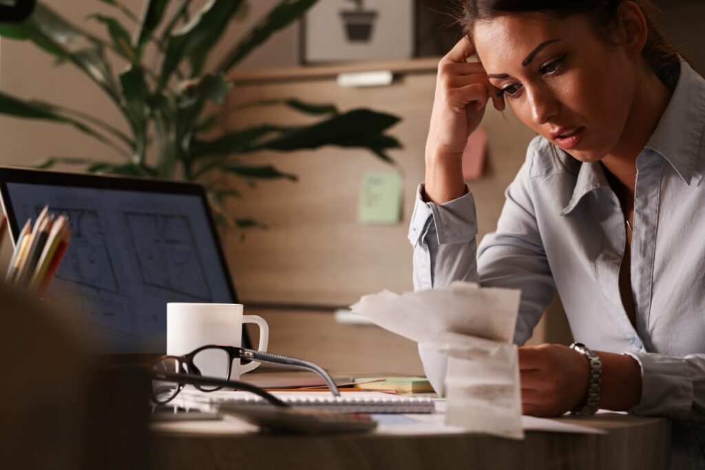Stressed woman reviewing bills and receipts at her desk while using a laptop, illustrating financial hardship and the challenges of managing debt before seeking debt relief options.
