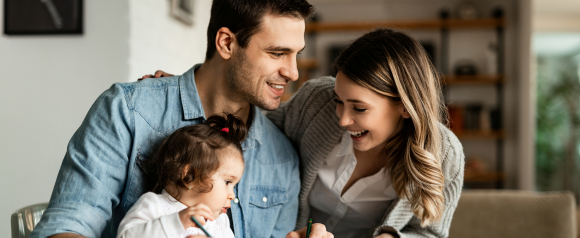 Parents smiling while sitting with their young daughter at a table as she draws. Warm family moment reflecting planning and protection with Fabric by Gerber Life life insurance.