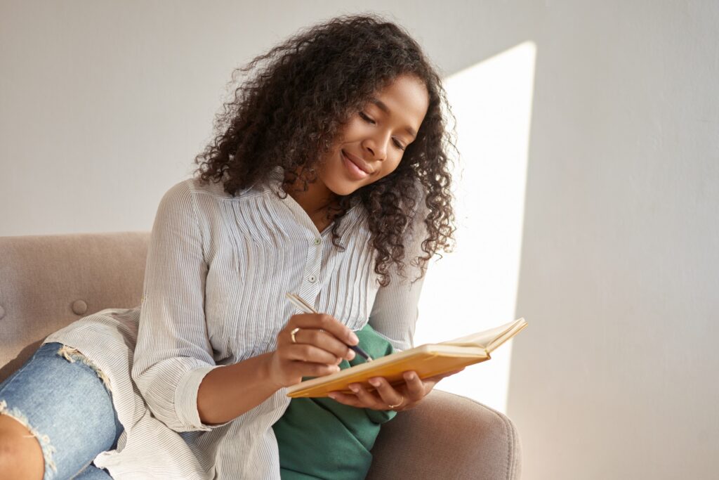 Woman sitting on a couch writing in a notebook, representing reflection, mindfulness, and learning practices related to mental wellness and consciousness books.