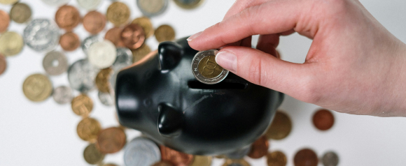 Hand placing a coin into a black piggy bank surrounded by scattered coins on a white surface. Visual representing saving money or planning finances with personal loans from City Bank.