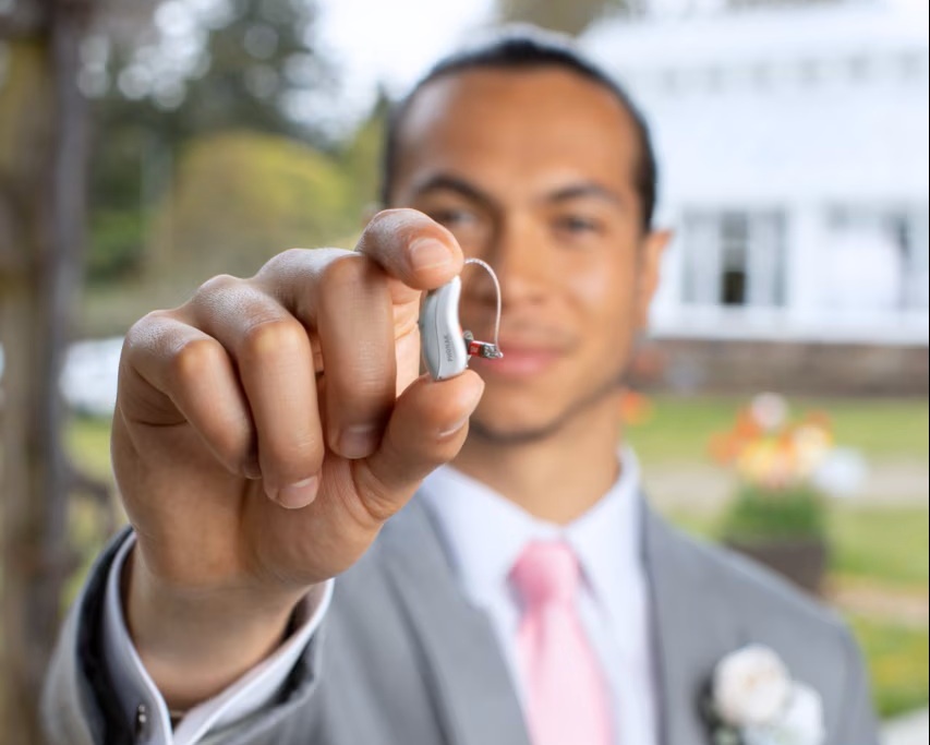 Man in a suit holding a small Phonak behind the ear hearing aid toward the camera while standing outdoors. The slim hearing aid with a thin clear wire and ear dome highlights modern hearing aids designed for discreet everyday use.
