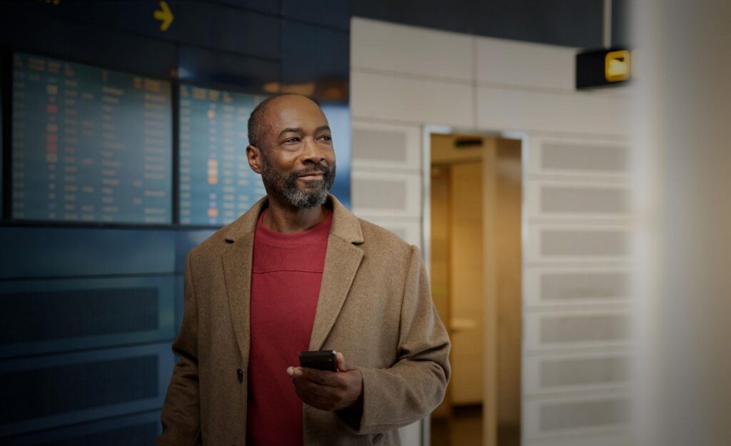 Man holding smartphone at airport terminal looking up at departure board, representing Auracast hearing aid public connectivity