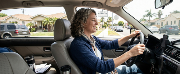 Smiling woman with curly hair driving through a sunny suburban neighborhood, relaxed behind the wheel