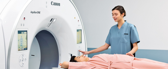 Medical technician in blue scrubs guiding a patient lying on a Canon Aquilion ONE CT scanner table in a modern radiology room