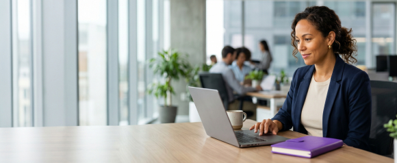 Professional woman in a navy blazer using a laptop at a desk in a bright modern open office with colleagues working in the background