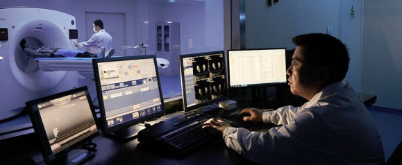 Radiologist operating multiple monitors in a darkened control room while a patient undergoes a CT scan in the adjacent imaging room