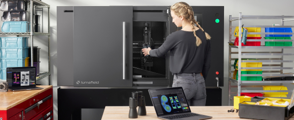 Technician loading a sample into a Lumafield Neptune CT scanner in an industrial lab with scan results visible on a nearby laptop