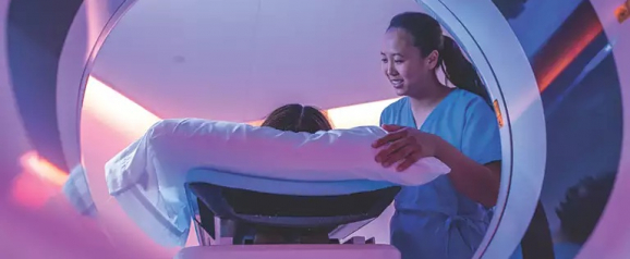 Medical technician in blue scrubs gently guiding a patient into a Philips CT scanner glowing with purple ambient lighting in a radiology suite