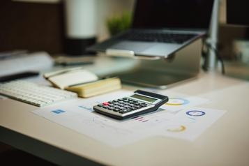 Calculator resting on financial charts next to a laptop and keyboard on an office desk.