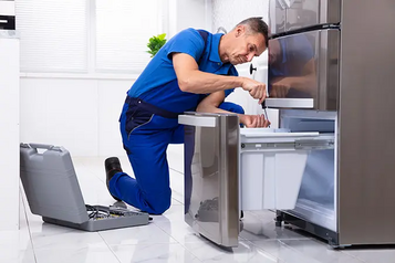 First American Home Warranty technician in blue uniform repairing a stainless steel refrigerator in a kitchen with a tool case open beside him