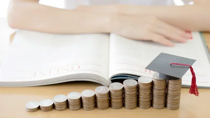 Rising stacks of coins topped with a graduation cap in front of an open book, illustrating the growing cost of student loans