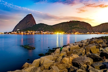 Sugarloaf Mountain and the Rio de Janeiro coastline at sunset, viewed across the bay from a rocky shoreline.