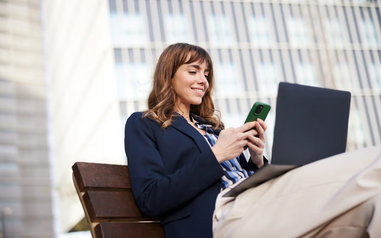 Smiling woman in business attire checking her phone with a laptop on her lap while sitting on a bench outside an office building